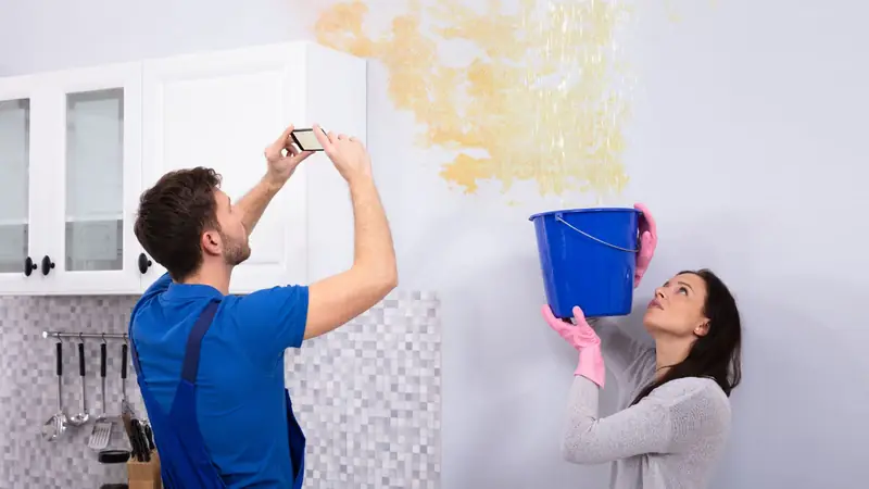 Man photographing water stained wall damage while a woman holds a bucket beneath the leak